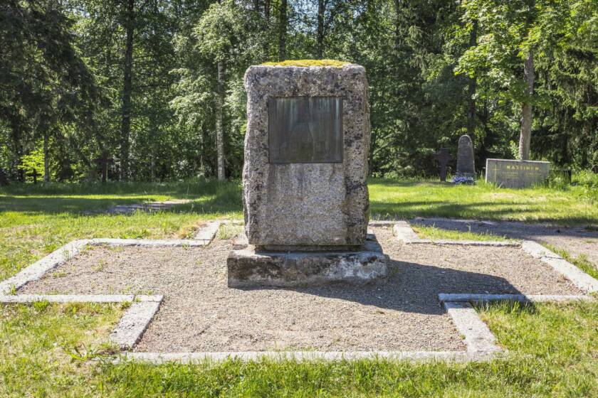 Kivinen pystypaasi, johon on kiinnitetty metallireliefi seisoo kivillä reunustetulla ristin muotoisella hiekka-alueella hautausmaalla.