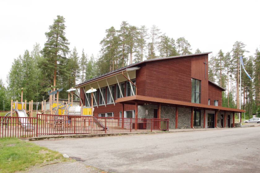 Kaksikerroksinen seurakuntakoti ja sen vieressä oleva lasten leikkikenttä auringonpaisteessa.