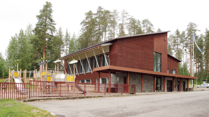 Kaksikerroksinen seurakuntakoti ja sen vieressä oleva lasten leikkikenttä auringonpaisteessa.