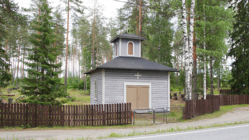 Tien vieressä pieni vaaleanharmaa ja hirsirakenteinen kappeli, jossa on pieni torni.