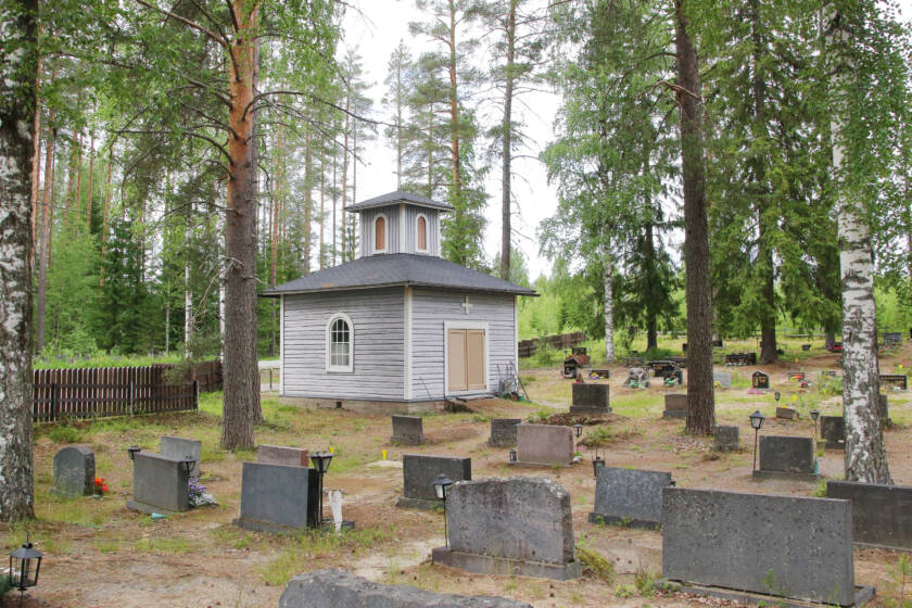 Vaalean pienen hirsirakenteisen kappelin vieressä on tummia hautakiviä.