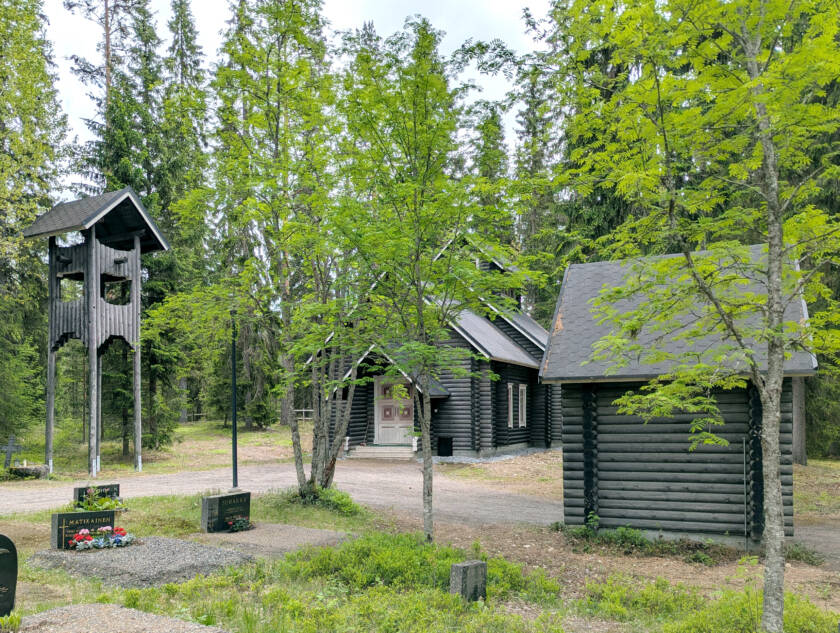 Hautausmaan vieressä harmaasta pyöröhirrestä tehdyt kellotorni, kappeli ja portti.