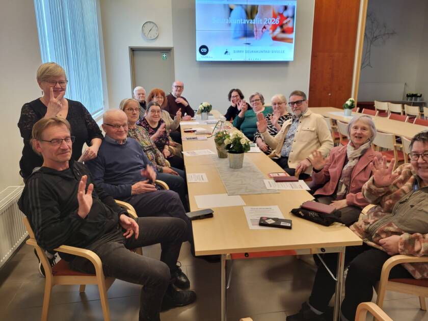 Pöydän äärellä istuvat vaali-illan osallistujat tervehtivät kuvaajaan päin.