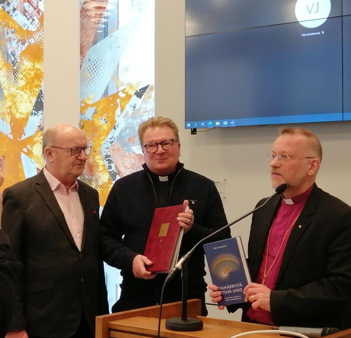 Kolme miestä seisoo, keskellä kirkkoherra pitää käsissään diplomia ja puhujapulpetin takana piispa esittelee kirjaa.