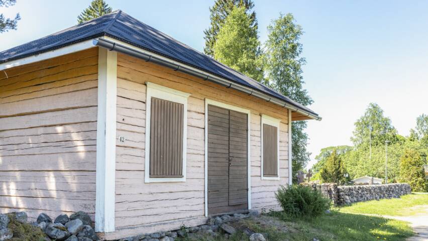 Vanhan pienen hirsirakennuksen ikkunat on suljettu luukuilla, samoin ovi on kiinni, rakennuksen vierellä on kiviaita ja vanhoja kuusia.