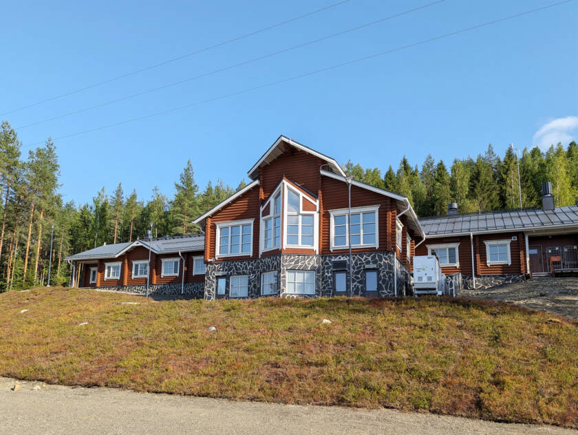 Mäntymetsän edessä, ruohikkoisessa rinteessä on pitkä hirsirakennus, jossa on isot ikkunat.