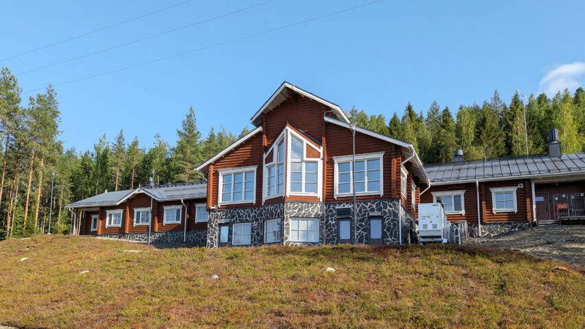 Mäntymetsän edessä, ruohikkoisessa rinteessä on pitkä hirsirakennus, jossa on isot ikkunat.
