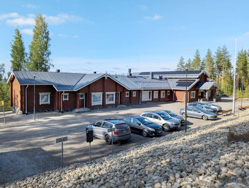 Kivikkoisen rinteen alla on autoja parkkipaikalla, niiden takana on pitkä hirsirakennus.