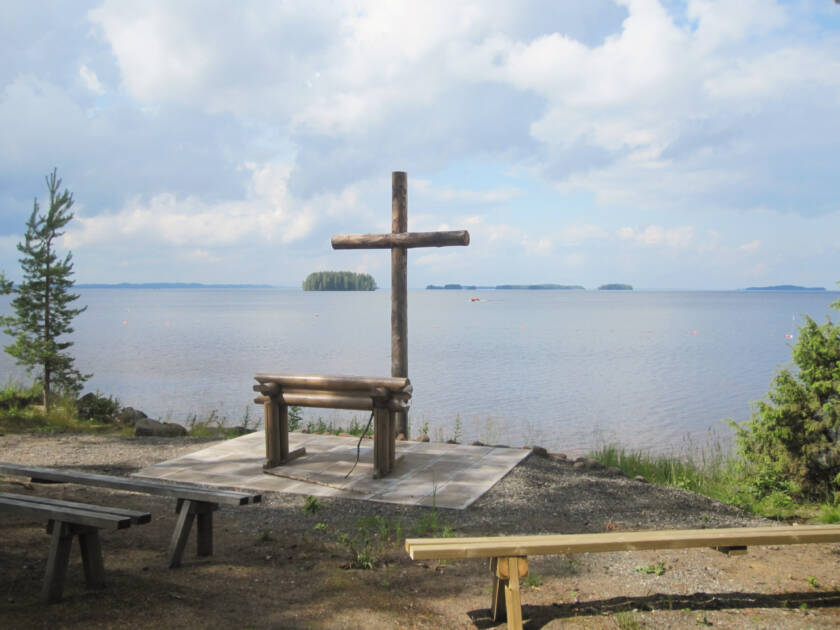 Kesäisen järven edessä rantahietikolla on suuri puuristi ja hirsinen alttaripöytä.