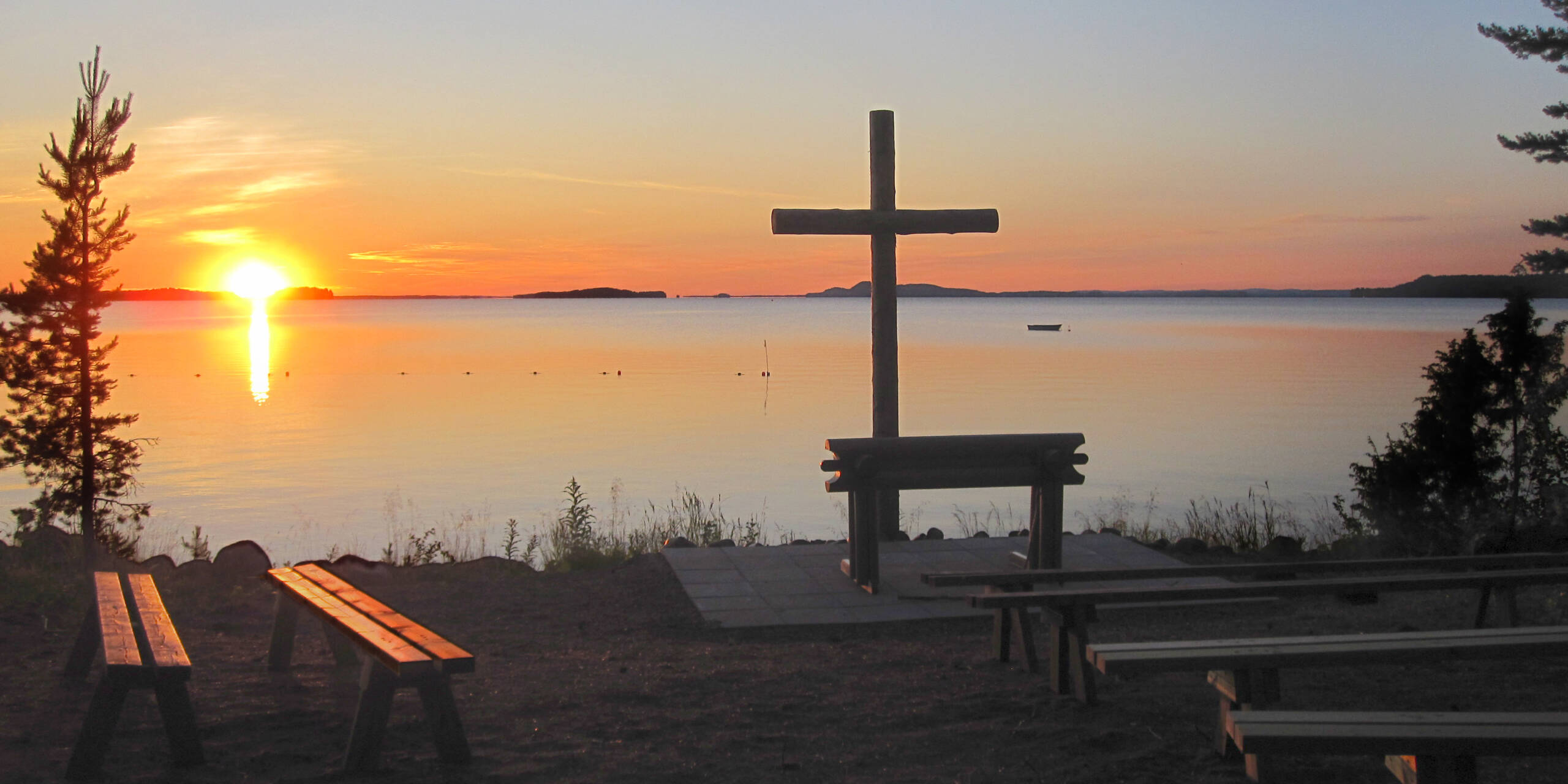 Aurinko laskee horisonttiin järven selän takana, etualalla rannalla on risti ja penkkejä.