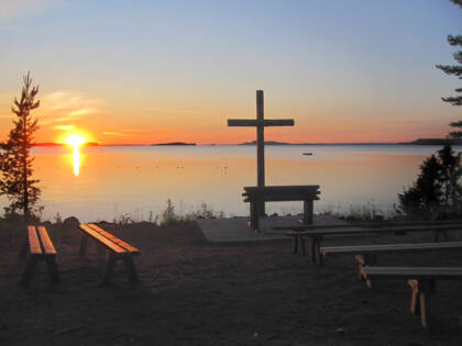 Aurinko laskee horisonttiin järven selän takana, etualalla rannalla on risti ja penkkejä.