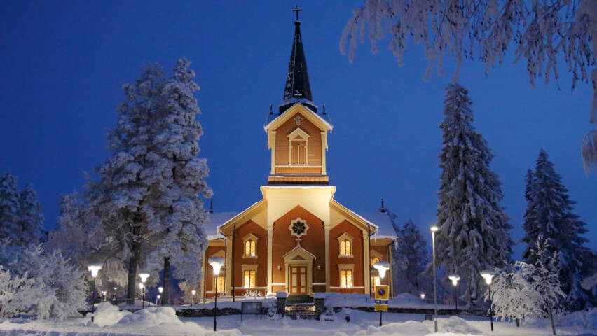 Puinen kirkkorakennus talvi-iltana, valot loistavat sen ikkunoista ja ympärillä maa ja puut ovat lumen peitossa.