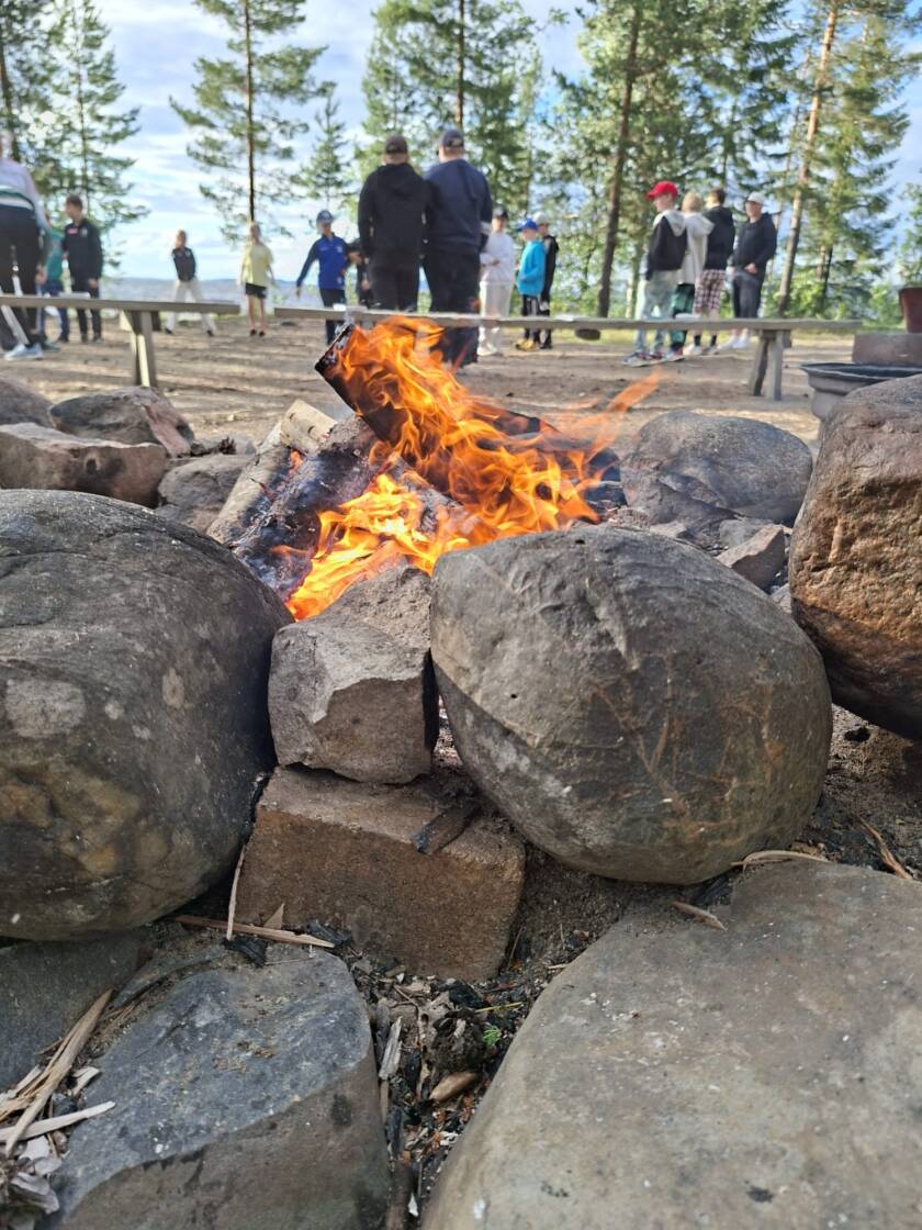 Isojen kivenlohkareiden lomassa on palaa nuotio, taaempana on puita ja seisovia nuoria.