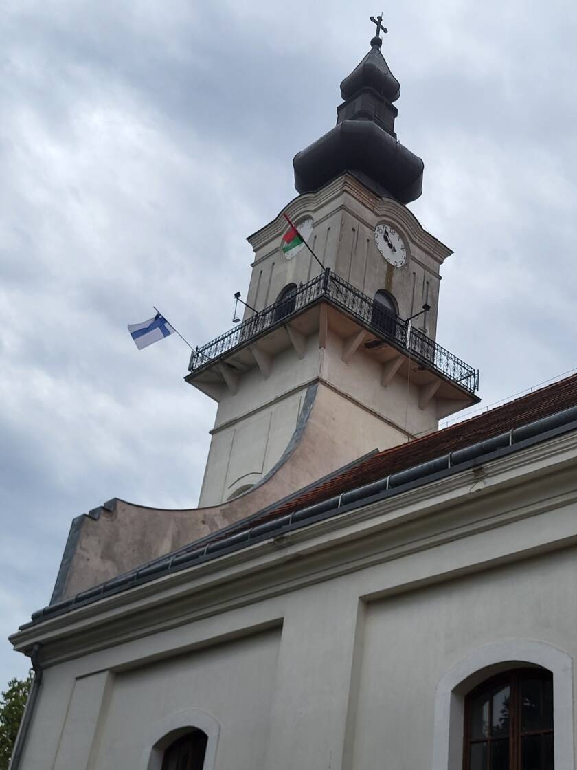 Taivas on pilvinen. Rakennuksen tornin ulkotasanteen koristeellisessa metallikaiteessa on kiinni kaksi lippua. Tasanteen yläpuolella on kello ja sipulinmuotoinen katto risteineen.