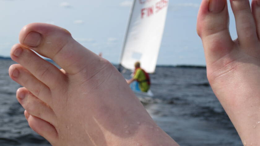 Henkilön varpaiden välistä näkyy järvellä kiitävä purjevene.