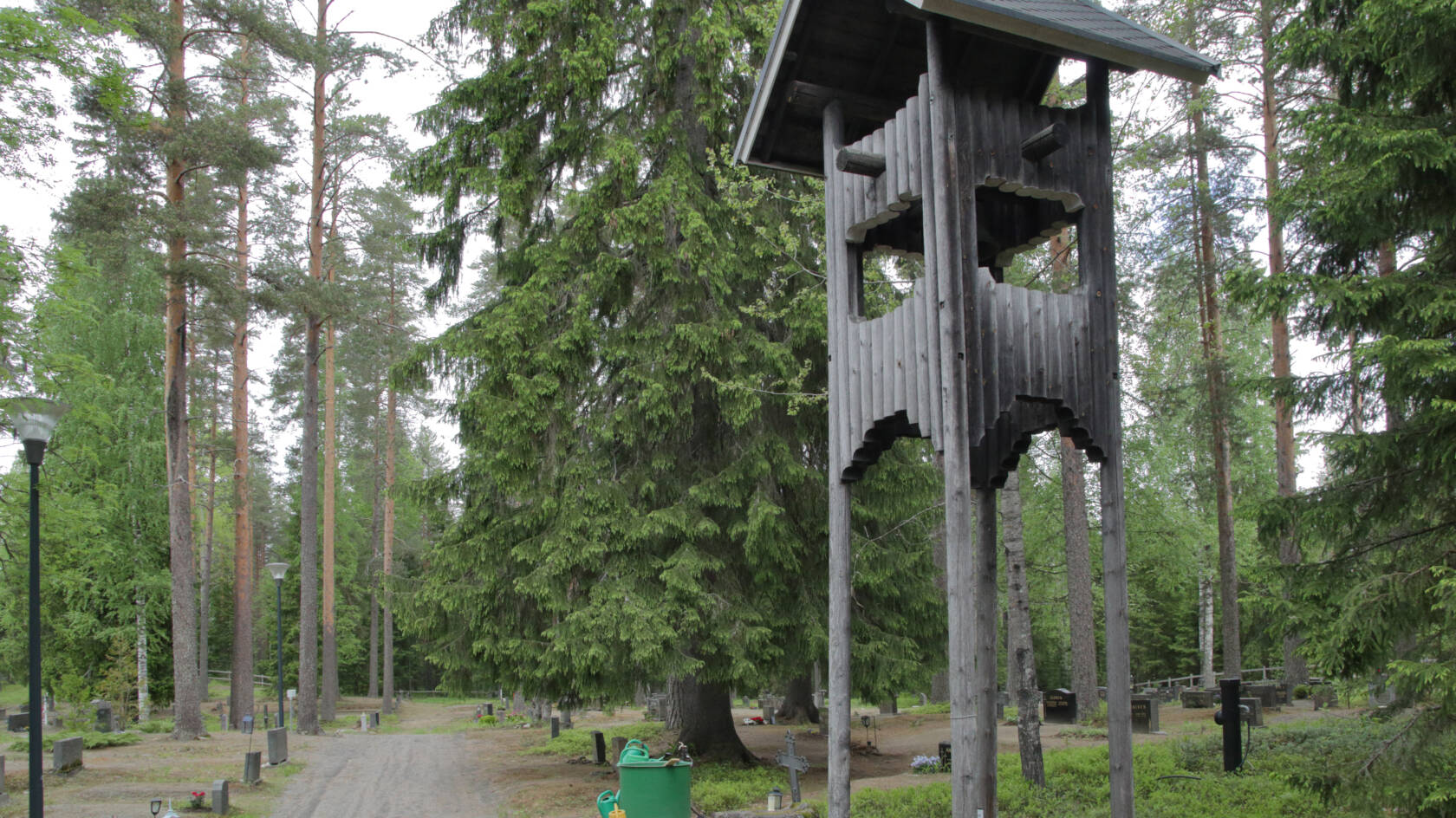 Isojen kuusten varjossa on hautakivia, etualalla harmaista pyöröhirsistä tehty kellotorni.