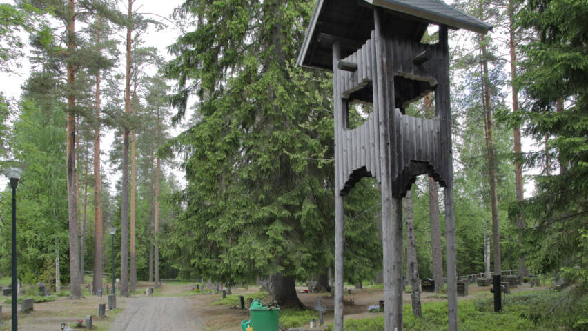 Isojen kuusten varjossa on hautakivia, etualalla harmaista pyöröhirsistä tehty kellotorni.