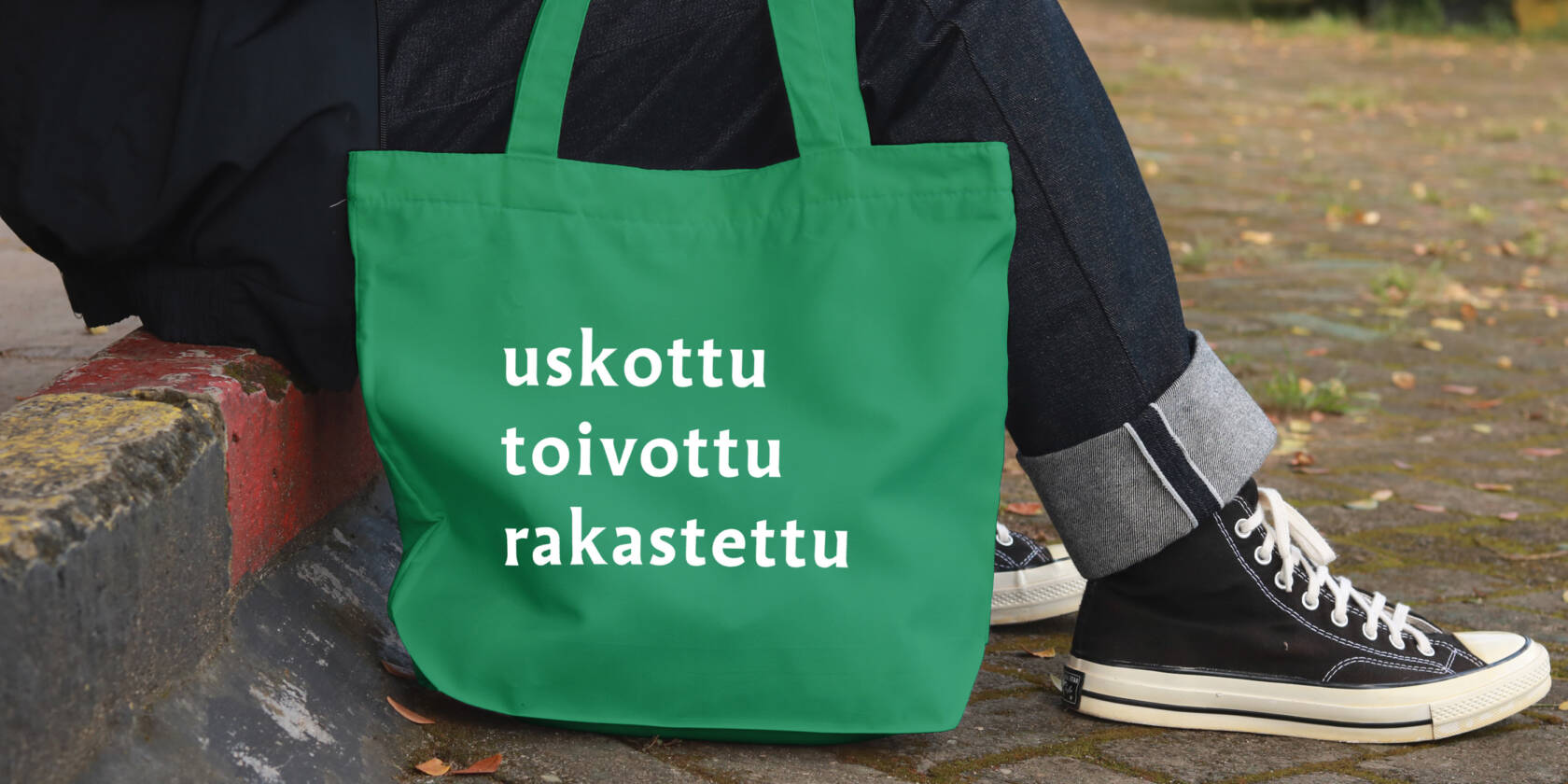 Jalkakäytävän reunalla istuvasta henkilöstä näkyvät vain jalat, vieressä on kangaskassi, johon on painettu sanat uskottu, toivottu, rakastettu.