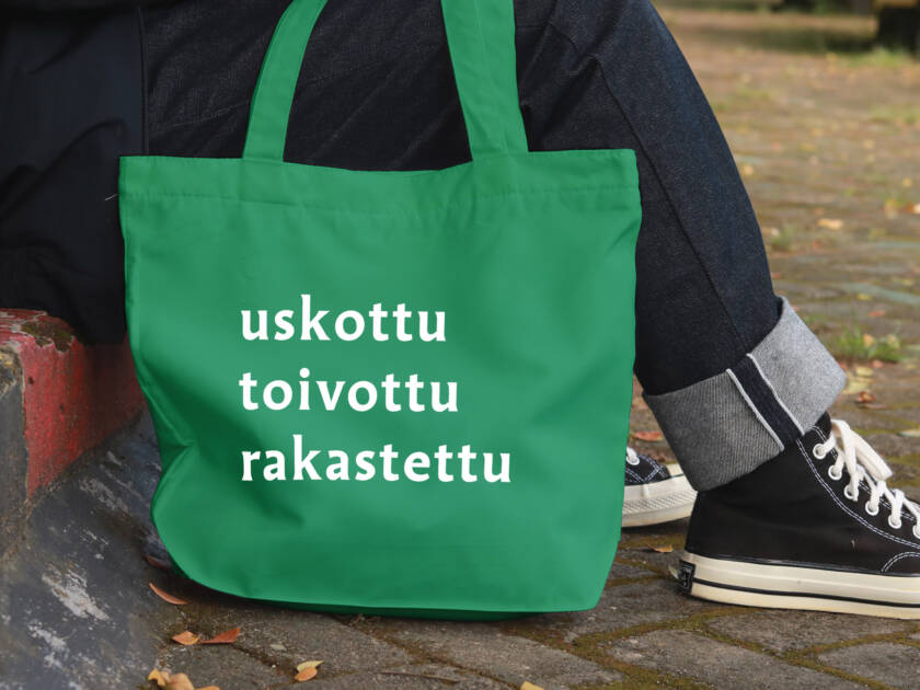Jalkakäytävän reunalla istuvasta henkilöstä näkyvät vain jalat, vieressä on kangaskassi, johon on painettu sanat uskottu, toivottu, rakastettu.