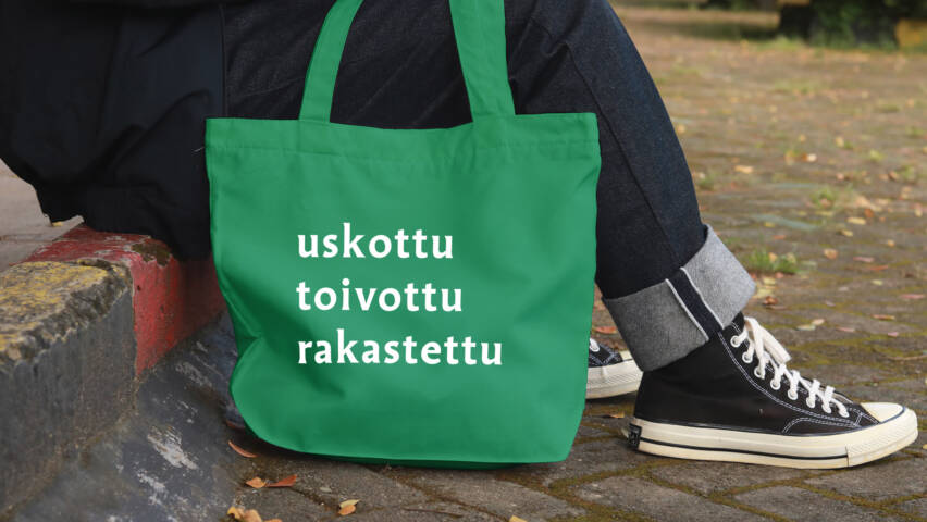 Jalkakäytävän reunalla istuvasta henkilöstä näkyvät vain jalat, vieressä on kangaskassi, johon on painettu sanat uskottu, toivottu, rakastettu.